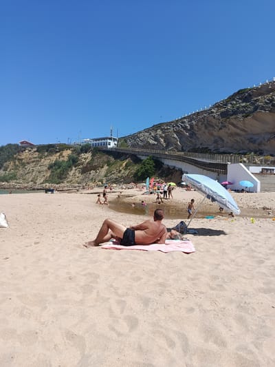 A wide shot shows a sandy beach with people sunbathing and playing in shallow water. In the foreground, a man is lying on a pink towel on the sand, facing away from the camera. He is wearing black swim trunks. Behind him, a striped beach umbrella provides shade. Further down the beach, other people are scattered around, some wading in a narrow stream of water that runs across the sand. In the background, a steep, rocky cliff rises up, with a building and a pedestrian walkway on top. The sky is a clear blue. This scene appears to be located at Praia da Aguda, near São João das Lampas, Portugal.