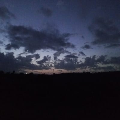 The image depicts a landscape scene at dusk or night, potentially in Ihururu, Kenya. The foreground is predominantly dark, obscuring the ground and any potential objects or subjects. Above the dark foreground is a horizon line, possibly indicating trees or vegetation. The sky occupies the upper portion of the image, filled with clouds varying in density and color. The clouds range from dark gray to lighter shades, indicating the remaining daylight or the presence of light sources. The overall impression is a dim or dark environment, suggesting either the onset of night or a heavily overcast day. There are no visible people or identifiable objects.