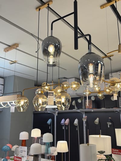 The image shows a retail display of various types of lighting fixtures, primarily chandeliers and lamps, in what appears to be a home goods or furniture store. The lighting fixtures vary in design and material, with some featuring clear glass globes with visible Edison-style bulbs, while others have opaque shades in white, pink, and blue.

The scene is indoors, likely a showroom, with shelves and a black wall as the backdrop. The lighting in the store is on, illuminating the products. The perspective is a mix of close-ups on individual fixtures and wider shots showing the arrangement of multiple items.

There are several price tags and product information labels visible. One label reads "Hadiah Langsung" which translates to "Instant Gift" in Indonesian. Another label says "Diskon" which means "Discount". There is also text that appears to be product names and prices, such as "Rp 999.000" and "Rp 199.000". The mention of "Pekalongan, Indonesia" suggests the location of the store.

The overall impression is a well-lit commercial space showcasing a variety of modern and decorative lighting options. No people are visible in the image, and the focus is entirely on the products. The time of day is not discernible.