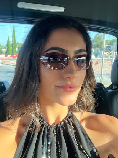 This is a candid, close-up, portrait-oriented photo of a woman with dark hair and light skin, wearing sunglasses and a black top adorned with sequins. The photo is taken from a low angle, with the woman's face tilted upwards. She is wearing large, oval-shaped sunglasses with dark lenses, and her lips are slightly parted, revealing a soft pink lipstick. Her hair is dark brown and wavy, falling around her face. The sun is casting a warm glow on her skin, highlighting her features and creating a slight sheen. The background is blurred, but appears to be the interior of a car, possibly looking out a window. The sunlight filtering through suggests it is daytime. There is no visible text in the image. The setting appears to be casual, given the selfie-style nature of the photo and the apparent vehicle interior. The overall mood is relaxed and perhaps a bit glamorous due to the sequined top and sunglasses.