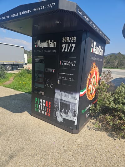 A black, freestanding automated pizza vending machine, branded "Le Napolitain - LA PIZZA TRADITION," is positioned outdoors on a light-colored, gravel-aggregate paved surface in Fréjus, France. The machine features a black canopy displaying "24H/24 • PIZZAS FRAÎCHES • 24H/24 • 7J/7" in white text. Its front panels advertise "24H/24 7J/7" and "CHAUDES EN 3 MINUTES," alongside "PIZZAS FRAÎCHES ARTISANALES" in red and green. An integrated touchscreen interface, a credit card reader, and a product dispensing slot are visible. Decorative elements include a prominent image of a cooked pizza and a vintage black-and-white photograph depicting several people at a table, labeled "PIZZERIA NAPOLETANA." Contact information, including a phone number and an email address, is also present. To the machine's left, green grass with yellow wildflowers and low shrubs borders the pavement. Behind this, a grey semi-trailer truck, partially visible and branded "KOOGEL," is parked on an asphalt lot. More green and brown shrubbery is present to the machine's right. In the background, distant trees and hills are discernible under a clear blue sky, suggesting a roadside rest area or commercial zone. No individuals are actively interacting with the machine.