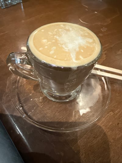 A close-up overhead shot shows a glass mug of coffee sitting on a clear glass saucer atop a wooden table. Two tan-colored stir sticks are partially visible behind the mug. The coffee has a light tan foamy top. The mug handle is clear glass. Light reflects off the glass and the polished table surface. The photo was taken during the day. It is possible this image was taken in Dania Beach, United States of America.