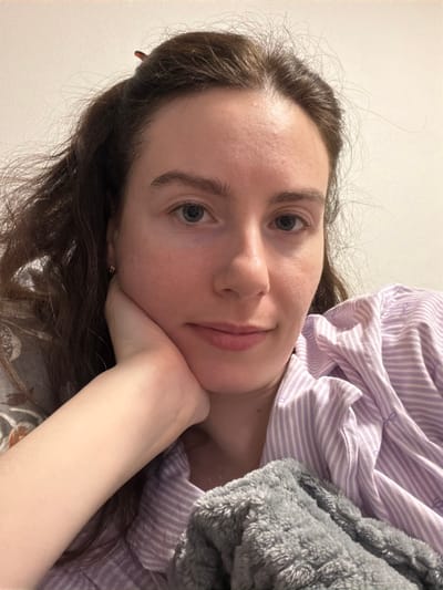 The image displays a close-up, head-and-shoulders portrait of a single female individual in an indoor setting. The individual has a pale complexion, light blue eyes, and dark brown, wavy hair that appears disheveled, with a dark hair clip visible at the crown of her head. She wears a light purple and white striped garment, possibly a pajama top, and a small metallic earring is visible in her left earlobe. Her left hand is positioned beneath her chin, partially supporting her head as she looks directly towards the camera with a subtle smile. A grey, textured, possibly faux fur blanket or throw covers her upper body on the right side of the frame. The background is a plain, light-colored wall. The specific location in the city of Ковалёво, Russia, cannot be determined from the visual information within the image.