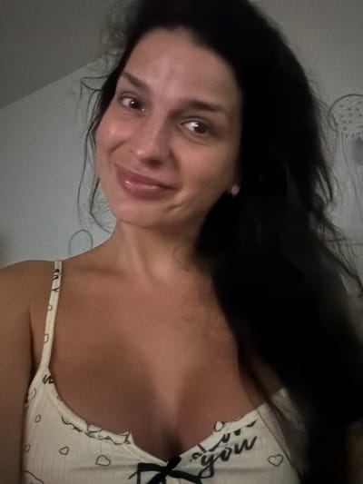 A woman with dark hair and brown eyes smiles at the camera. She is wearing a white camisole with small heart patterns and the words "love you" printed on it. The background appears to be a wall in a room. The image is a selfie taken from the chest up. There are no details in the image that suggest a location in Lauderdale Lakes, United States of America.