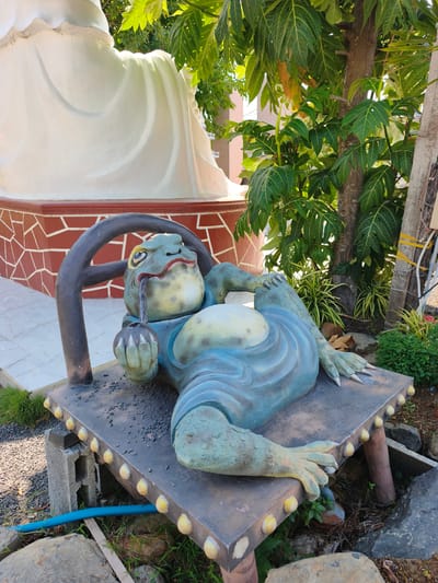 A statue of a frog is depicted in the foreground of the image, lying on a bench. The frog statue is green and blue and is holding a smoking pipe. In the background, a large white statue is visible, along with trees and foliage. The scene is outdoors, with a tiled wall behind the statues. This image was taken in Tuy An Nam, Vietnam.
