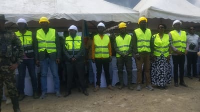 The image shows a group of 11 people standing in front of a white tent in Sake, Democratic Republic of Congo. The people are dressed in formal and casual clothing, with some wearing hard hats and yellow safety vests. One person is wearing military fatigues. They are standing in front of the tent, with some of the people standing behind chairs. The ground is dirt. The location is a public area, most likely outside. The people are likely at a construction site or other public gathering.