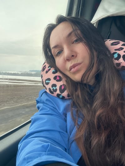 A young woman with long, wavy brown hair is resting her head on a pink and black leopard-print neck pillow. She is wearing a light blue long-sleeved shirt. She has fair skin, light-colored eyes, and slightly parted lips.

The background shows a landscape visible through a window, possibly a car window. The landscape appears to be a flat, snowy field. The sky is overcast and gray.

Based on the prompt request, the location is Bagrash-Bigra, Russia. However, there is no way to forensically verify this location from the image alone.