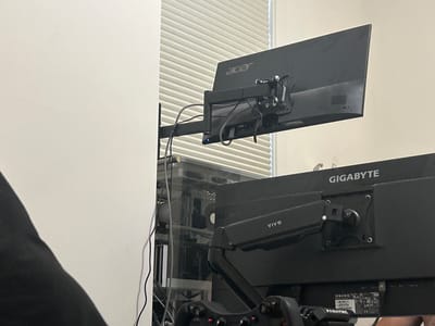The image shows a close-up, eye-level view of two computer monitors mounted on adjustable arms. The monitor in the foreground is a black Gigabyte monitor, partially obscured by its arm. The monitor behind it is a black Acer monitor. Both monitors are mounted on black, articulated monitor arms. The monitors are attached to a vertical stand, which is not fully visible. In the background, a computer tower with a transparent side panel is visible, revealing internal components. A black piece of fabric, possibly clothing, is in the lower left corner, partially obscuring the scene. A window with horizontal blinds is visible behind the Acer monitor. The overall scene suggests a home office or gaming setup. There are no people visible in the image. The location of Richmond Hill, Canada, is not ascertainable from the visual information presented in the image.