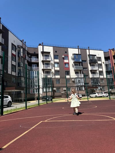 A young girl with blonde hair, wearing a light green dress and white sneakers, is running on a red outdoor sports court. She is positioned in the center of the frame, facing away from the camera. The court has yellow line markings. Behind the court, a green chain-link fence separates it from a paved area with parked cars, including a white SUV and two white sedans. Further in the background, a multi-story modern apartment building with a brown and white facade, and balconies, dominates the scene. The sky is clear and blue. The setting appears to be a residential courtyard in the city of Vyazniki, Russia.