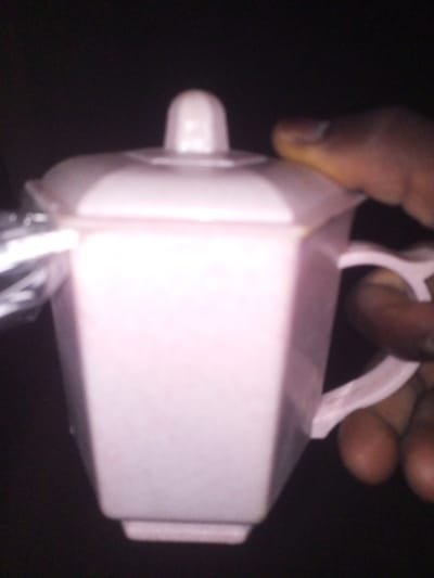 A person holds a pink, polygonal-shaped container with a lid. The container has a small, round handle on the right side and a lid with a knob on top. It's held at an angle, with the person's fingers visible gripping the handle. The surrounding background is dark and out of focus. A shiny metallic object is near the top left of the container.