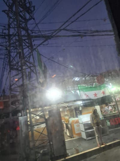 A bustling street scene is captured through a window, reflecting tangled electrical wires against a deep blue sky. Buildings line the street, adorned with signs and a prominent Syrian flag. A person is visible near a storefront. The scene is illuminated by a bright light, possibly a street lamp, which creates a strong glare. The image appears to be taken at dusk or early evening.