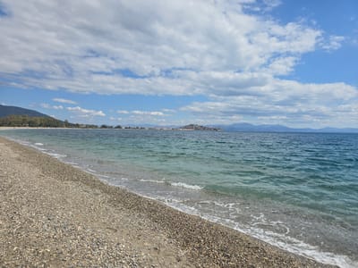 The image depicts a scenic view of a pebble beach in Astros, Greece. The foreground is dominated by the textured surface of the beach, covered with various sizes and colors of pebbles. Gentle waves lap at the shore, creating a thin line of white foam. The sea is a clear, turquoise hue, stretching towards the horizon. In the mid-ground, a distant coastline is visible, with a cluster of buildings perched on a hill, suggesting a small town or village. Beyond the coastline, a range of mountains forms a hazy silhouette against the sky. The sky itself is a vibrant blue, dotted with a mix of white, fluffy clouds and larger, more dispersed formations. The overall impression is one of a tranquil, natural setting with a touch of coastal habitation.