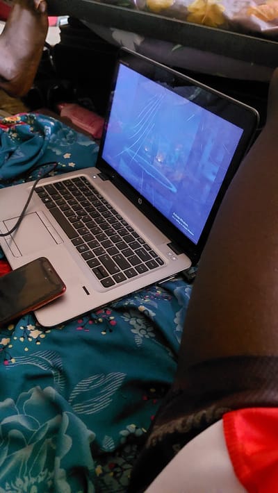 A low-angle, close-up shot captures a laptop on a bed with teal, floral patterned sheets. The laptop screen is cracked and displays a blue background with a "Activate Windows" prompt. A red-cased smartphone rests to the left of the laptop, partially covering the touchpad. The scene appears to be indoors, possibly in a bedroom, in New Nyanya, Nigeria. The presence of a foot in the upper left corner suggests a person is present but not fully visible. The overall lighting is dim, contributing to a relaxed or casual atmosphere.