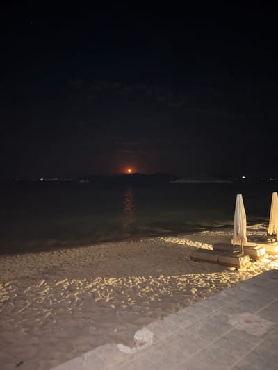 An evening view of a sandy beach in Nea Peramos, Greece, shows the moon rising over the sea. The moon, an orange orb, is partially obscured by a distant landmass and casts a shimmering reflection on the water. In the background, faint lights suggest a coastal town or settlement. The foreground features beach chairs and closed umbrellas, indicating the end of the day. The sand is illuminated by artificial lighting, creating distinct shadows. The overall scene captures a tranquil night at the seaside.