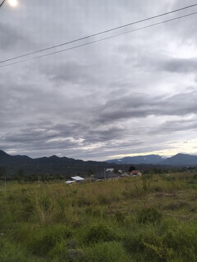 This is a photograph of a cloudy, overcast day in Panteraya, Indonesia. The scene is a rural landscape featuring rolling hills and mountains in the distance, partially obscured by clouds. In the foreground, there is a field of tall, dry grass, some of which appears to be golden and straw-like. Scattered within the field and further back are a few buildings, some with visible white or orange roofs. The sky is a uniform gray with thick, horizontal cloud cover. Two thin power lines are visible stretching across the upper portion of the image. The overall mood is subdued and somewhat somber, likely due to the weather conditions. The time of day is difficult to ascertain precisely, but the dim lighting suggests it is either early morning or late afternoon. There is no visible text in the image.
