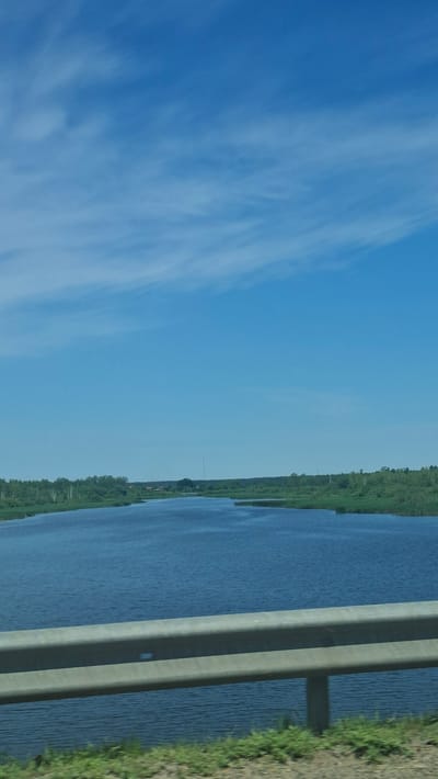 This media file depicts a landscape scene, likely taken from a moving vehicle, as indicated by the horizontal orientation of the photograph and the blurred vegetation on the right. The primary subjects are a wide body of water on the right side of the frame, and a vast expanse of blue sky with wispy white clouds occupying the left side. A concrete barrier, likely a bridge railing, runs along the right edge of the water, separating it from a strip of dry, grassy embankment.
The water appears calm, with subtle ripples on its surface. Along the far bank of the water, a dense line of green trees and vegetation stretches horizontally across the mid-ground, suggesting a rural or natural setting. A few small buildings and what appears to be a radio tower are faintly visible amidst the trees, hinting at a distant settlement. The sky is clear and bright, with soft, elongated clouds streaking across it.
The overall impression is of a serene, daytime scene in a natural environment. There are no visible people or specific objects of interest beyond the natural elements and the man-made barrier. The lighting suggests it is daytime, possibly late morning or afternoon, given the brightness of the sky. The location context of Драчкаўскі сельскі Савет, Belarus, implies a landscape typical of that region, likely a river or lake setting. No text is visible in the image.