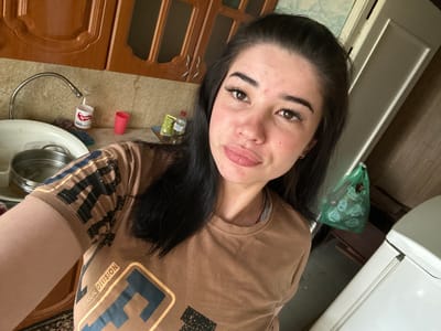 In a domestic kitchen setting, a young woman with long dark hair and brown eyes takes a selfie. She is wearing a brown t-shirt with a graphic print that includes the words "CASUAL DIVISION." The kitchen features wooden cabinets above a tiled backsplash and a stainless steel sink with a gooseneck faucet. On the counter, there are various items, including a pink cup, a white container with red lettering, a sponge, a blue dish with a bottle of water and a packet of chips, and a metal colander in the sink. To the right of the woman, a white refrigerator is partially visible, and a green plastic bag hangs on a wooden wall. The overall impression is a candid moment captured in a home environment.