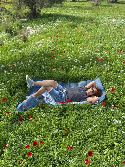 The image displays a single individual resting on a blue mat within a vibrant green field abundant with red and white wildflowers, characteristic of spring in the Modiin-Maccabim-Reut area of Israel. The person is lying supine, with their head positioned towards the right side of the frame. They are observed wearing a horizontally striped t-shirt, denim shorts, and light grey athletic shoes. Sunglasses obscure the eyes, and reddish-brown hair is visible around the face. Their hands are clasped behind their head, and legs are slightly bent and crossed. A striped towel is partially visible beneath the individual's torso, and a denim bag or folded garment rests near their lower legs on the mat. The background features sparse trees and exposed rocks under clear daylight conditions.