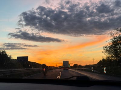 The image depicts a view from inside a moving vehicle, likely a car, driving on a highway. The location appears to be near Vakarel, Bulgaria, based on the roadside landscape and the style of chevron markers visible on the right shoulder. The sky is filled with a vibrant sunset, featuring shades of orange and streaks of cirrus clouds. Darker, more dense clouds are present towards the top of the frame. Traffic is visible on the highway ahead, including a motorcycle in the left lane and a truck in the center lane. There are roadside signs and vegetation on either side of the highway. Water droplets are visible on the windshield. The interior of the car, including the dashboard and steering wheel, is visible in the foreground. A cross can be spotted in the distance on the right side of the road.