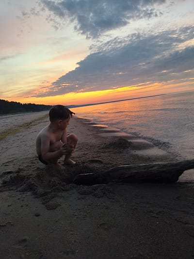 A young boy is crouching on a sandy beach at sunset in Apšuciems, Latvia. The sky is filled with colorful clouds, with hues of orange, yellow, and grey. The ocean is calm, reflecting the colors of the sunset. A large piece of driftwood lies in the foreground on the sand. The boy appears to be playing in the sand, holding some in his hands. The overall scene is peaceful and serene, capturing a moment of childhood on a beautiful summer evening.