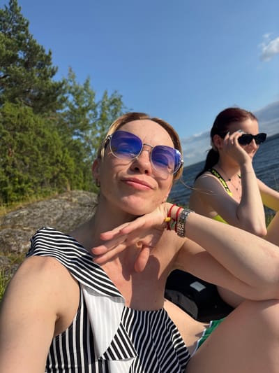 Two women are seated outdoors, on a rocky shoreline, by a body of water under a clear blue sky. The woman in the foreground, on the left, is blonde and wearing a black and white striped swimsuit and blue sunglasses. She is resting her chin on her hand and smiling. She is also wearing several bracelets on her wrist. The woman in the background, on the right, has dark hair and is wearing a yellow swimsuit and black sunglasses. She is looking towards the left and appears to be talking on her phone. Behind the women, trees and a rocky embankment can be seen. The water is visible in the background, with a slight ripple on its surface. The overall scene suggests a casual, sunny day by the water. The location is identified as Shuiskaya Chupa, Russia.
