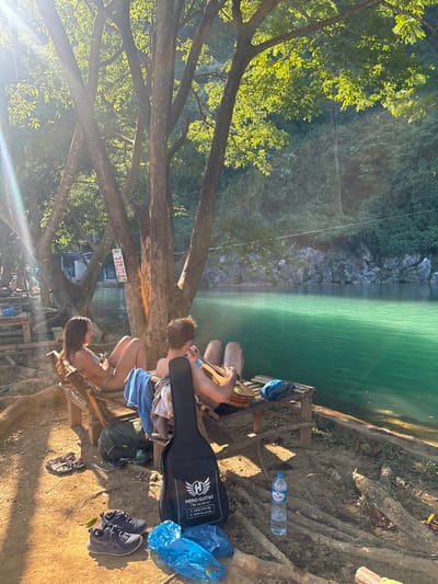 A couple relaxes by a turquoise lake. The man plays guitar while sitting on a bench beside the woman. A guitar case, water bottle, and shoes are on the ground nearby. Lush green foliage and tree branches surround them, with sunlight filtering through the leaves. The landscape transitions to rock formations on the far side of the lake.