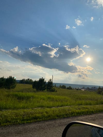 The image captures a rural landscape in the vicinity of Novoye Selo, Russia, under a dynamic sky. A bright sun is partially obscured by a large, dramatic cloud formation, casting visible sunbeams downwards. The foreground consists of a gravel road and a vibrant green grassy field dotted with several small coniferous trees. In the middle ground, a gently sloping hill is covered in grass and sparse trees, with a few buildings visible in the distance, suggesting a small settlement. The background features a hazy horizon line of distant hills and trees. A utility pole and overhead wires are present on the right side of the image. The overall scene is a daytime outdoor view, possibly captured from a vehicle given the reflection in the lower right corner, which appears to be a side-view mirror.
