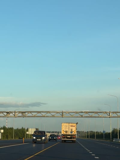 The image depicts a view from a moving vehicle on a multi-lane highway, likely in the vicinity of Kaipy, Russia, given the context. The sky is a clear, pale blue, with some faint clouds visible near the horizon. A large metal overhead gantry, used for traffic signs or lighting, spans the highway. On the right side of the highway, a row of streetlights stretches into the distance. The road surface is asphalt, with clear yellow and white lane markings.

In the foreground, a white truck with a box trailer is traveling in the right lane. The truck appears to be a commercial vehicle, possibly for logistics or delivery, with some markings on its trailer that are partially obscured or difficult to discern. To the left of the truck, a dark-colored van or minivan is also traveling in the right lane, followed by other vehicles. Further down the highway, a black sedan is visible in the left lane. On the left side of the highway, a dense line of green trees forms a backdrop, with a few white road signs placed intermittently. The overall scene suggests daytime traffic on a highway.