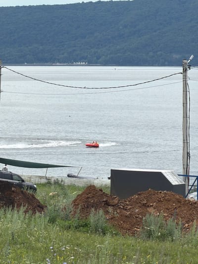 In Sergeevka, Russia, a wide body of water, possibly a river or a lake, stretches towards a densely forested hill. The water's surface is choppy, with visible wakes indicating movement. In the middle distance, a red inflatable tube, carrying two people, is being pulled across the water, leaving a white spray. The foreground of the image is dominated by a grassy area with scattered wildflowers and two large mounds of dark soil. A dark-colored SUV is parked to the left, partially obscured by a green tarp stretched over a frame. To the right, a gray, angular structure sits atop another pile of soil, with a blue metal fence behind it. Electrical poles and wires are visible in the mid-ground, crossing the frame horizontally and vertically. The sky is overcast, contributing to the muted lighting of the scene.