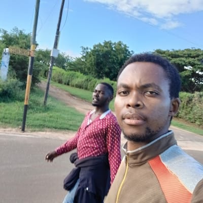 The image depicts two adult males outdoors on Ocean Drive in Tanga, Tanzania. In the foreground, positioned to the right, a dark-complexioned male with short, dark hair and a beard is visible from the chest up, seemingly capturing a selfie. He wears a brown zip-up jacket featuring orange and light-colored striped panels on the shoulders and chest. His expression is neutral.
Behind and to the left of the foreground subject, a second dark-complexioned male walks on the paved road, partially out of focus. He wears a long-sleeved maroon shirt with white dotted patterns and a white collar, with a dark jacket tied around his waist. His mouth is slightly open, and his gaze is directed towards the left side of the frame.
To the left, a black and yellow striped utility pole bears a yellow street sign with black lettering, clearly reading "OCEAN DRIVE." Another utility pole is visible further behind it. A dirt path ascends uphill from the main paved road, bordered by green grass and lush trees in the background. The sky above is clear blue with scattered white clouds. The overall environment suggests a daytime scene in a verdant, semi-urban or suburban area.