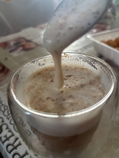 A close-up, low-angle shot shows a clear glass mug with a double wall, filled with a frothy, light brown beverage, likely coffee or hot chocolate, being poured from above. The beverage has a foamy top with small brown flecks, possibly cinnamon or coffee grounds. The mug rests on a patterned tablecloth. In the background, a white plastic container with some golden-brown food items, possibly pastries or cookies, is visible. The image is taken indoors, possibly in a home or cafe setting, with soft, natural lighting. The scene does not provide any specific information or context about the city of Zubov, Russia.