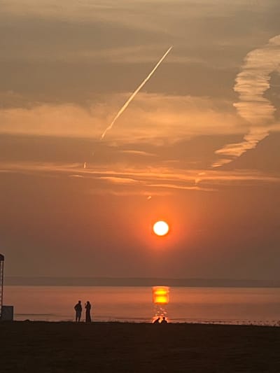 The image captures a sunset over a body of water, possibly a sea or large lake, with a distinct contrail from an aircraft stretching across the sky. The sun, a bright orange orb, is positioned in the lower center of the frame, casting a warm glow and creating a reflection on the water's surface that appears to be rectangular in shape. In the foreground, silhouetted against the water, are two figures standing on what appears to be a beach or shoreline. The landscape is generally flat, with the horizon line clearly defined. The sky is filled with wispy clouds that are illuminated by the setting sun. The lower portion of the image shows a dark foreground, likely land, with some texture suggesting sand or grass. The scene conveys a sense of tranquility and natural beauty. While the location is stated as Surakovo, Russia, there are no specific landmarks visible in the image that would definitively confirm this.