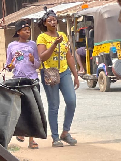 The image depicts a daytime urban street scene in Neni, Nigeria. Two females are walking on a dusty ground surface. The individual on the left, appearing younger, wears a black baseball cap backward, a light purple long-sleeved shirt with a "SPRINKLE make my AWESOME" cupcake graphic, and dark flip-flops. She holds a yellow and a red object in her right hand. The individual on the right wears a black headwrap, a yellow short-sleeved t-shirt with cartoon character designs, blue skinny jeans, and dark Crocs-style clogs. She carries a small, brown patterned cross-body bag with a gold-colored clasp. Her right hand is raised to her mouth, holding a small light-colored object, and her mouth is open as if speaking or reacting. In the mid-ground to the right, a black and yellow three-wheeled auto-rickshaw (Keke Napep) with blue accents and an orange dome light is visible. A person's bare leg is visible within the vehicle. Building structures with corrugated metal roofs are partially visible in the background. A large, black, soft-sided container hangs from a metal frame in the immediate foreground, partially obscuring the lower left portion of the scene.