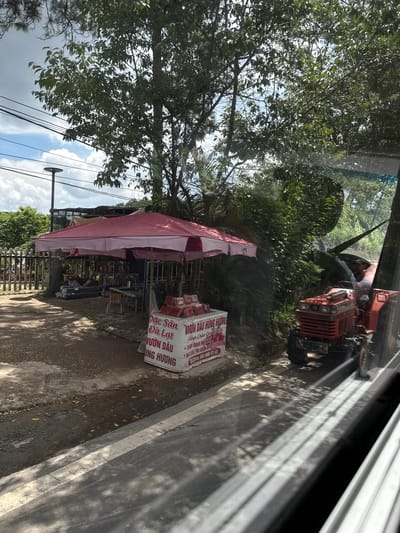 In Thôn 6, Vietnam, a roadside stall with a pink and white striped awning displays various goods, including boxes of what appear to be strawberries and other packaged items. Behind the stall, stacked rolls of dark material are visible. A red tractor, driven by a person wearing a hat, is parked to the right of the stall on the dirt road. The scene is bordered by a wooden fence and lush green foliage, with a cloudy sky overhead. A street lamp is also visible in the background. The image appears to be taken from inside a vehicle, as evidenced by the reflections on the glass.