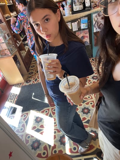 A woman in Fredericksburg, Virginia, USA, holds two iced coffee drinks at the Agora Downtown Coffee Shop. The shop's interior features shelves with items for sale, as well as decorative tile flooring. The woman has long brown hair, is wearing a dark blue shirt and jeans and has yellow painted nails. A second woman with long brown hair is partially visible at the top of the image.
