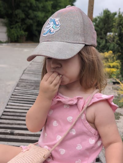 A young child, likely a toddler, is seated outdoors, wearing a pink dress adorned with white hearts and a sparkling silver baseball cap featuring a unicorn patch. The child is bringing a small, light-colored object to their mouth with their right hand, suggesting they are eating. They are positioned on a wooden bench with a slatted surface, which appears to be part of an outdoor seating area. The background consists of lush green trees and foliage, with hints of yellow flowers. The setting suggests a park or a garden, possibly in Perliavka, Ukraine, given the context. The child also wears a small, woven pink purse with a braided strap.