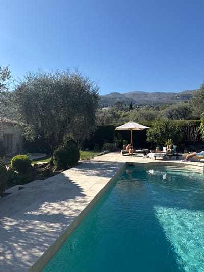 The image shows a sunlit swimming pool area in what appears to be a private villa. Located in Tourrettes-sur-Loup, France, the area features the pool situated next to a stone-tiled patio. Lounge chairs are present, some occupied by people. An umbrella provides shade. The pool area overlooks a lush garden with mature trees and manicured shrubbery. Distant mountains are visible under a clear blue sky.
