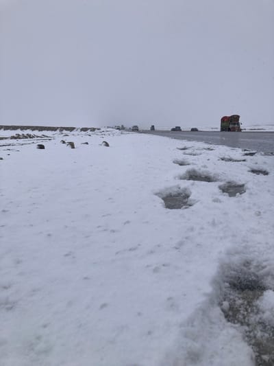 This image captures a snow-covered landscape alongside a road in Khanai Baba, Pakistan, under an overcast sky. In the foreground, a layer of fresh white snow covers the ground, featuring distinct human footprints leading towards the road on the right. Several dark, irregular rocks are partially visible within the snow on the left. To the right, the edge of an asphalt road is visible, appearing wet or icy. On the road, multiple vehicles are present, moving away from the viewer. Most prominent is a large, multi-axle truck on the right side, heavily loaded with cargo secured under green and red tarpaulins. Further along the road, a line of approximately six to seven additional vehicles, including cars and smaller trucks, can be seen. The sky is uniformly light gray and overcast, with visible snowfall or precipitation in the air. A low, snow-covered embankment or ridge runs parallel to the road on the left side, separating the immediate foreground from the distant, hazy horizon.