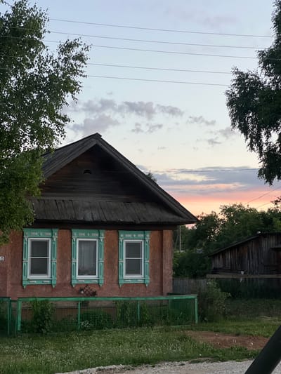This is a photograph of a weathered, wooden house with red-brown stucco walls, located in the village of Pigozino, Russia. Three windows with ornate, teal-colored frames and shutters are visible on the front facade. A low, green metal fence encloses a small yard with overgrown grass and wildflowers. The roof of the house is made of dark, aged wood. In the background, a darker wooden structure, possibly a shed or another house, is visible, along with lush green trees and a hint of a pastel-colored sunset in the sky. Overhead, power lines stretch across the frame. No people or animals are present in the image.