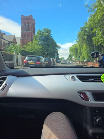 The image is taken from inside a car, looking out through the windshield on a sunny day. The foreground shows the dashboard of the car, with a silver trim and a black textured surface. A person's leg, clad in beige, patterned trousers, is visible in the lower right corner.

Outside the car, a street scene unfolds. A red brick church tower with arched windows and a flagpole is prominent in the background, on the left side of the image. Adjacent to the church are residential buildings and trees. The street is lined with trees that create a canopy over the road, and there are other cars visible, suggesting traffic. Traffic lights are visible, indicating a road intersection ahead. The sky is blue with some scattered clouds. The overall impression is a view from a car driving through a residential area of Stockton-on-Tees, United Kingdom, on a bright, clear day.