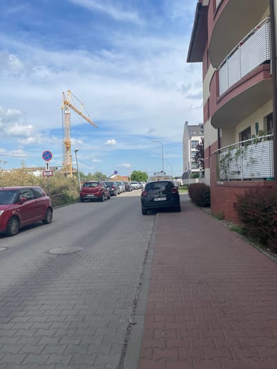 The image shows a street in Wysoka, Poland. Several cars are parked along the side of the road, including a black Citroen with license plate DMR 7834U and a red Suzuki. A construction crane is visible in the background, under a blue sky. A street sign is visible. The road is paved with gray and brown pavers. The buildings are modern with balconies.