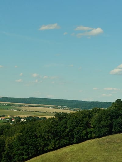 This is a medium-shot, eye-level, outdoor, daylight, landscape photograph taken in the city of Șcheia, Romania. The scene depicts a rolling green hill in the foreground, covered with grass. Beyond the hill, a dense forest of green trees stretches across the middle ground. In the background, a small village with red-roofed houses is nestled amidst agricultural fields of varying shades of green and yellow, with a line of dark green trees forming a backdrop to the settlement. The sky above is a clear, bright blue with a few scattered white clouds. The overall impression is one of a serene rural landscape on a sunny day.