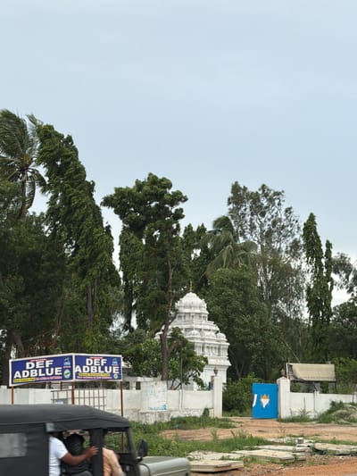 The image shows a street scene in Thimmapur, India, under an overcast sky. On the left, a sign with blue lettering reads "DEF ADBLUE" and "Diesel Exhaust Fluid". To the right of the sign, a white temple with intricate carvings is visible, surrounded by lush green trees. A blue gate with a white emblem is positioned in front of a white wall. In the foreground, a three-wheeled vehicle, possibly an auto-rickshaw, is partially visible, with two people inside. The ground is covered in dirt and vegetation, with some concrete slabs scattered about.