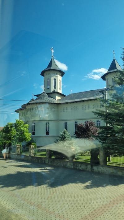 The image shows a large, light-colored building, likely a church or monastery, with two prominent towers topped with crosses. The building has a gray, sloped roof and a cream-colored facade with arched windows. In front of the building is a paved walkway with a decorative fence. Several trees, including evergreens and deciduous trees, are visible around the property. The sky is blue with a few scattered clouds. A person in a white top and dark pants is walking on the paved area to the left of the frame. The scene appears to be in a town or rural area, with hints of other buildings and vegetation in the background. The location is identified as Milișăuți, Romania.