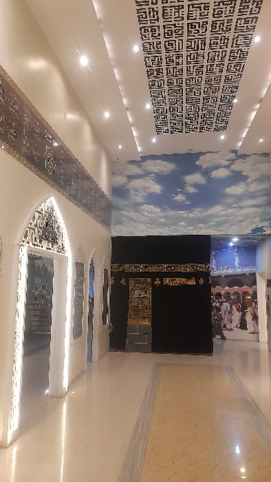 The image depicts an indoor exhibition space or cultural center located in Glantengan, Indonesia. The scene is well-lit by artificial light, primarily from ceiling-mounted recessed spotlights and backlighting. The floor consists of highly polished light-colored tiles, with a central pathway distinguished by a light brown, veined section bordered by darker grey strips. The ceiling is white, featuring a prominent rectangular panel with intricate black Kufic calligraphy or geometric Islamic patterns.
On the left side of the room, a white wall displays a decorative frieze with intricate patterns running horizontally near the ceiling. Below this, multiple arched openings are present. The closest arch on the left is ornately decorated with laser-cut or carved Islamic patterns and is brightly backlit, revealing a staircase beyond it. Other similar arched elements are partially visible further along the wall.
In the center and right portion of the image, a large-scale replica of the Kaaba, a cubic black structure with gold-colored decorative elements, including a golden door and a band of Arabic calligraphy (Kiswah), stands prominently. Above this replica, a large mural or wallpaper depicts a bright blue sky filled with white cumulus clouds. To the right of the Kaaba replica, another large mural depicts a crowd of individuals dressed in white pilgrim attire, indicative of Hajj pilgrims, gathered in a large courtyard, with a building resembling the Grand Mosque and possibly the Abraj Al-Bait clock tower in the background. No live individuals are present in the immediate foreground of the scene.