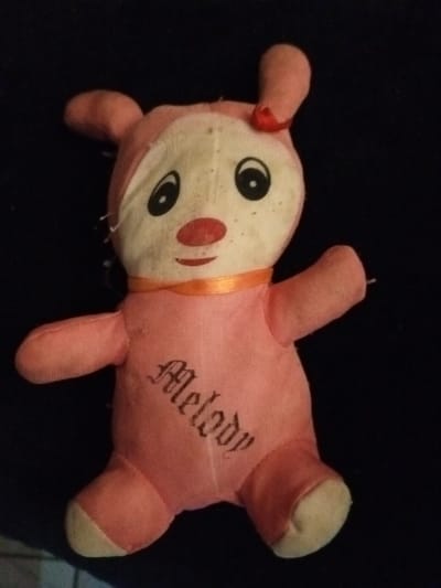 A worn, pink and cream stuffed animal resembling a rabbit sits against a dark background. It features a light-colored face with large, black eyes, a red nose, and a pink body. The rabbit has floppy ears, one adorned with a small red bow. A thin orange ribbon encircles its neck. The name "Melody" is stitched vertically in gothic font on its stomach. The stuffed animal appears aged and slightly damaged, with exposed stitching and faded colors.