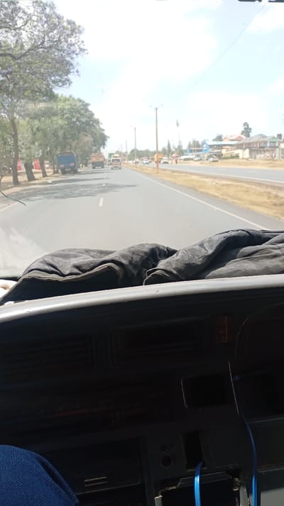 A road stretches ahead, bordered by trees and sparse buildings. Multiple vehicles are in the distance. The viewpoint appears from inside a vehicle, with the dashboard and a dark cloth prominent in the foreground. Blue cables are visible near the bottom right. The scene is captured during daylight hours.