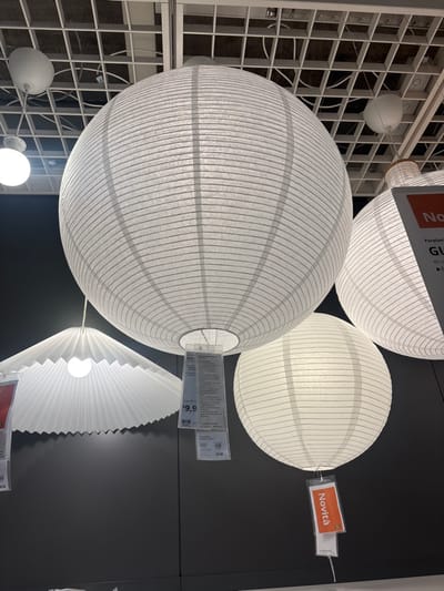 The image displays a retail lighting section, specifically showcasing multiple white paper lampshades and light fixtures suspended from a white grid ceiling structure. The prominent central object is a large, spherical, ribbed white paper lantern. To its right, a similar but partially cropped white ribbed paper lantern is visible, featuring a tag with "Novità" (Italian for 'New') and "Paralum..." (likely 'Paralume' for lampshade) printed on it. Below the central lantern, a smaller, fully spherical white ribbed paper lantern hangs, also adorned with an orange "Novità" tag. To the left and slightly below the central lantern, a distinctive fanned and pleated white paper lampshade with a wavy bottom edge is displayed. A transparent price tag, bearing the IKEA logo, the text "GUL," and a price of "€ 9,9," is attached to the central large lantern. Several smaller, white, dome-shaped light fixtures are integrated into the overhead ceiling grid. The background behind the hanging fixtures is a dark, solid-colored display wall. No individuals are present. The scene is located within an IKEA store in Corsico, Italy.