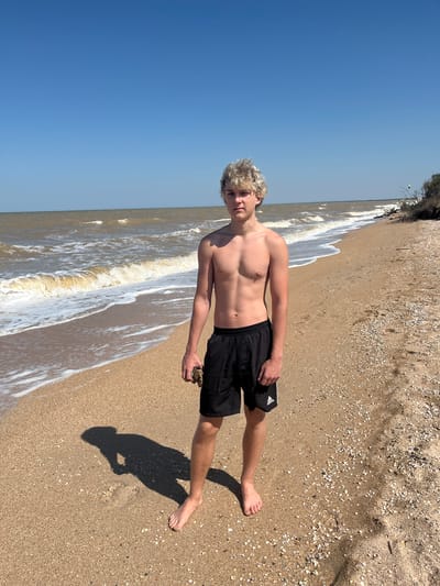 A young Caucasian male with blond, curly hair stands on a sandy beach, facing the camera. He is shirtless and wearing black Adidas shorts. His right hand is holding a small piece of driftwood. The ocean is to his left, with moderate waves breaking on the shore. The sky is clear and blue. The beach is made of sand mixed with small shells. In the distance, a small green kite can be seen in the sky, suggesting activities like kitesurfing. The location is identified as Dolzhanskaya, Russia. The overall scene depicts a sunny day at the beach.