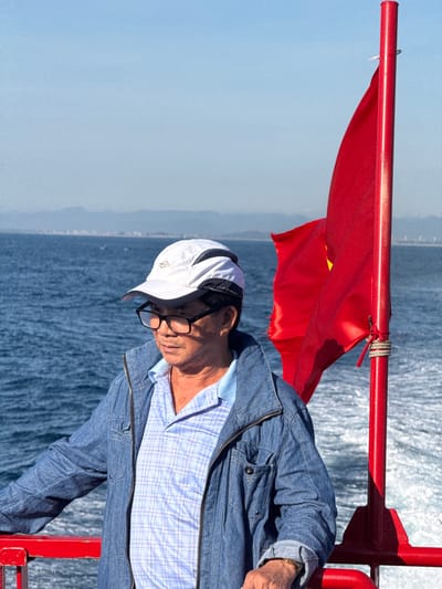 A man wearing glasses, a white and black baseball cap, and a blue denim jacket over a plaid collared shirt stands on a boat. A red flag with a yellow star, likely the Vietnamese flag, is attached to a red pole to his right. The boat is moving through the water, creating a wake. In the background, distant mountains and a hazy coastline are visible. The sky is clear and blue. The scene appears to be located off the coast of An Hải, Vietnam.